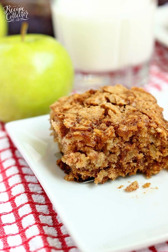 a square of golden brown cake with streusel topping on a white plate with a green apple in the background